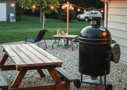 Robust barbecue smoker on gravel patio with smoke rising, weathered picnic table nearby under string lights, and hanging grilling utensils, creating a cozy outdoor gathering atmosphere.