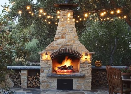 Outdoor kitchen fireplace with brick pizza oven in a rustic patio, surrounded by weathered stones and twinkling string lights, creating a cozy atmosphere for al fresco dining.