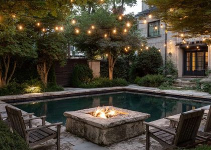 Tranquil backyard scene featuring a square stone fire pit beside an inground pool, surrounded by lush greenery and illuminated by overhead string lights, creating a cozy oasis for relaxation and gatherings.